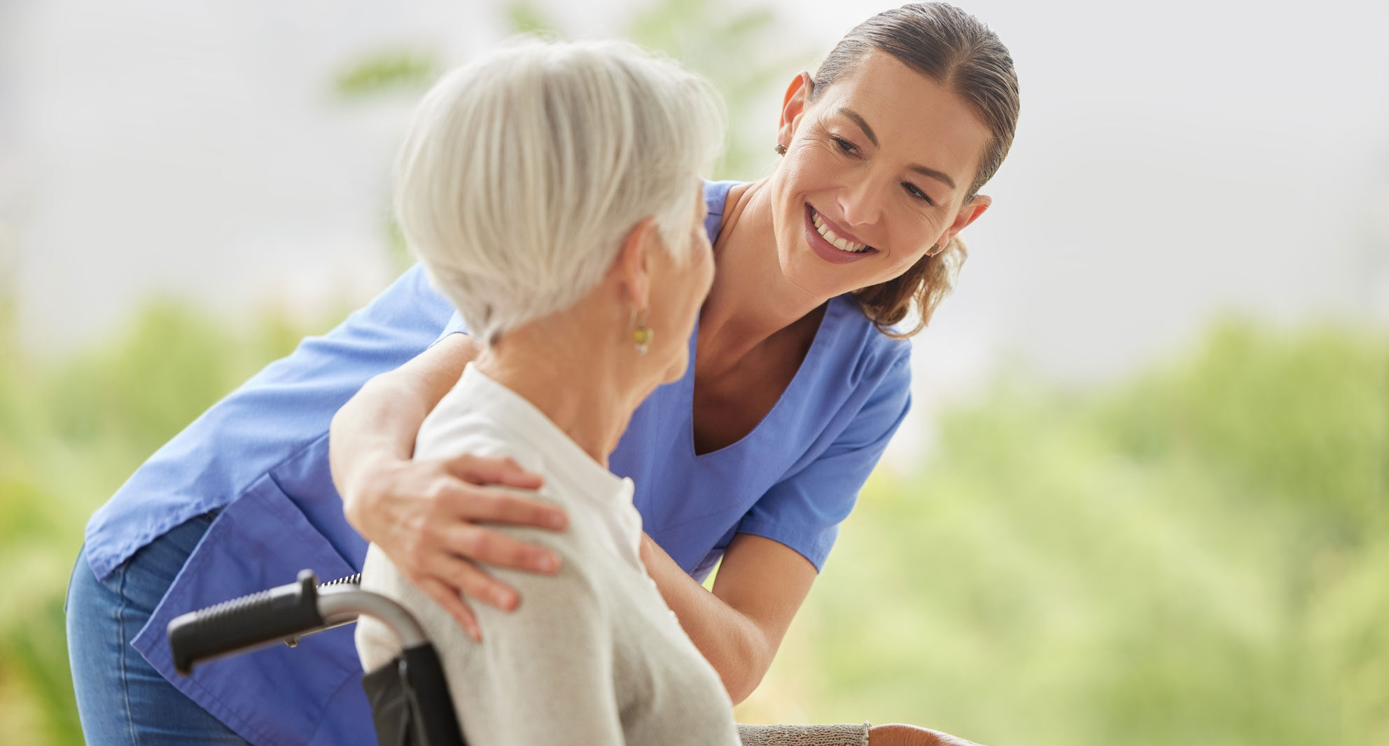 Personal de cuidados paliativos con bata azul sosteniendo los hombros y sonriendo a un anciano en silla de ruedas