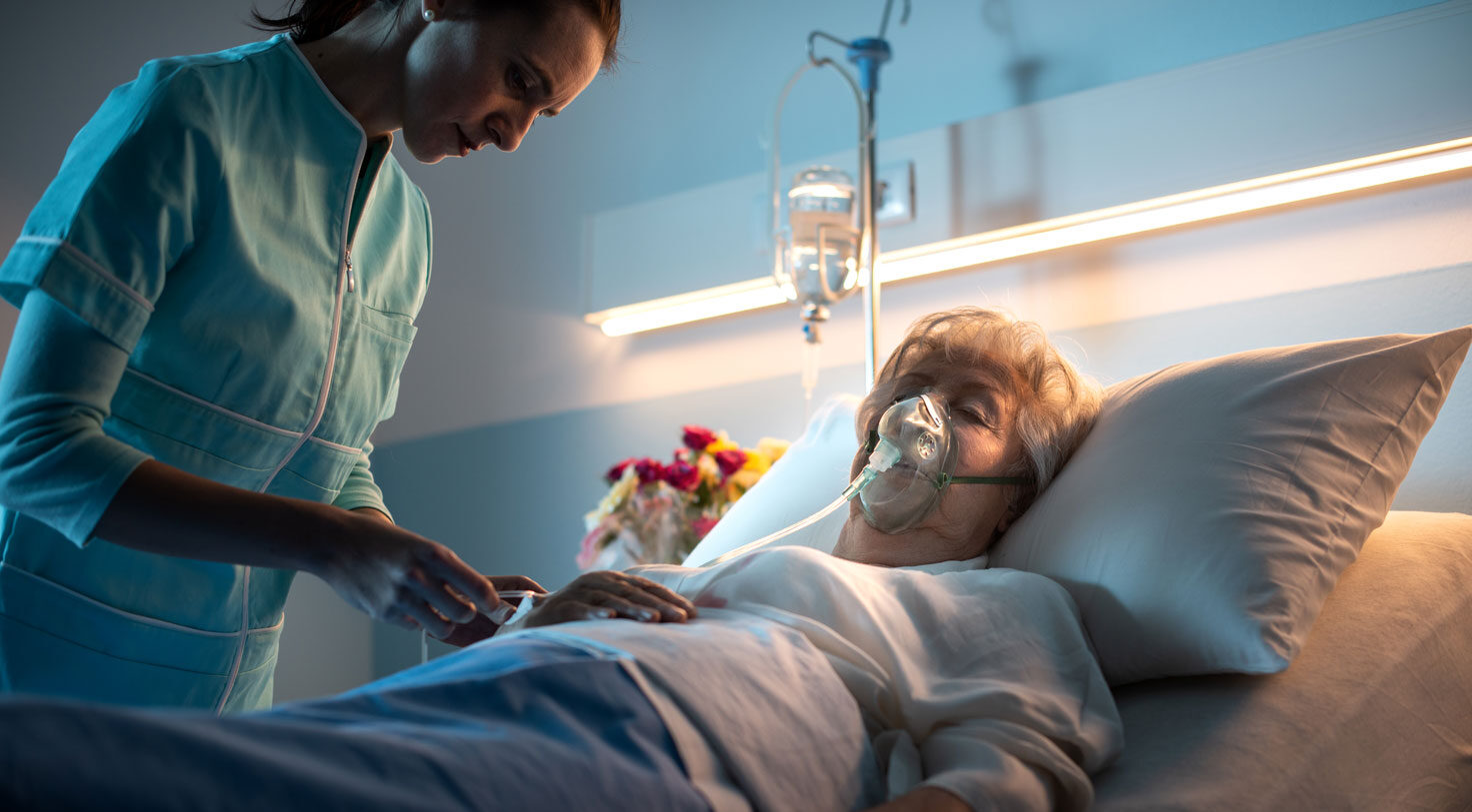 Enfermera de cuidados paliativos con bata azul revisando a un paciente anciano postrado en cama inconsciente con una máscara de oxígeno