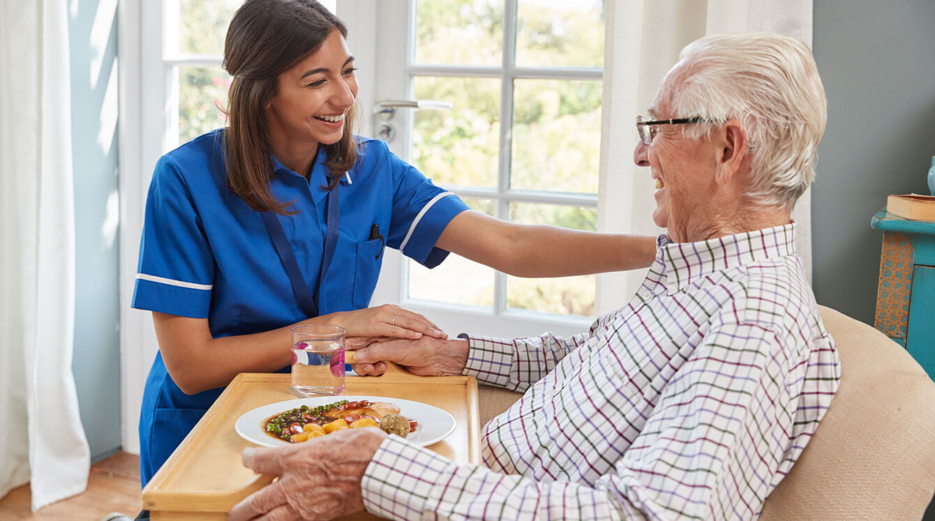 A caregiver in blue scrubs assisting an elderly gentleman about to eat
