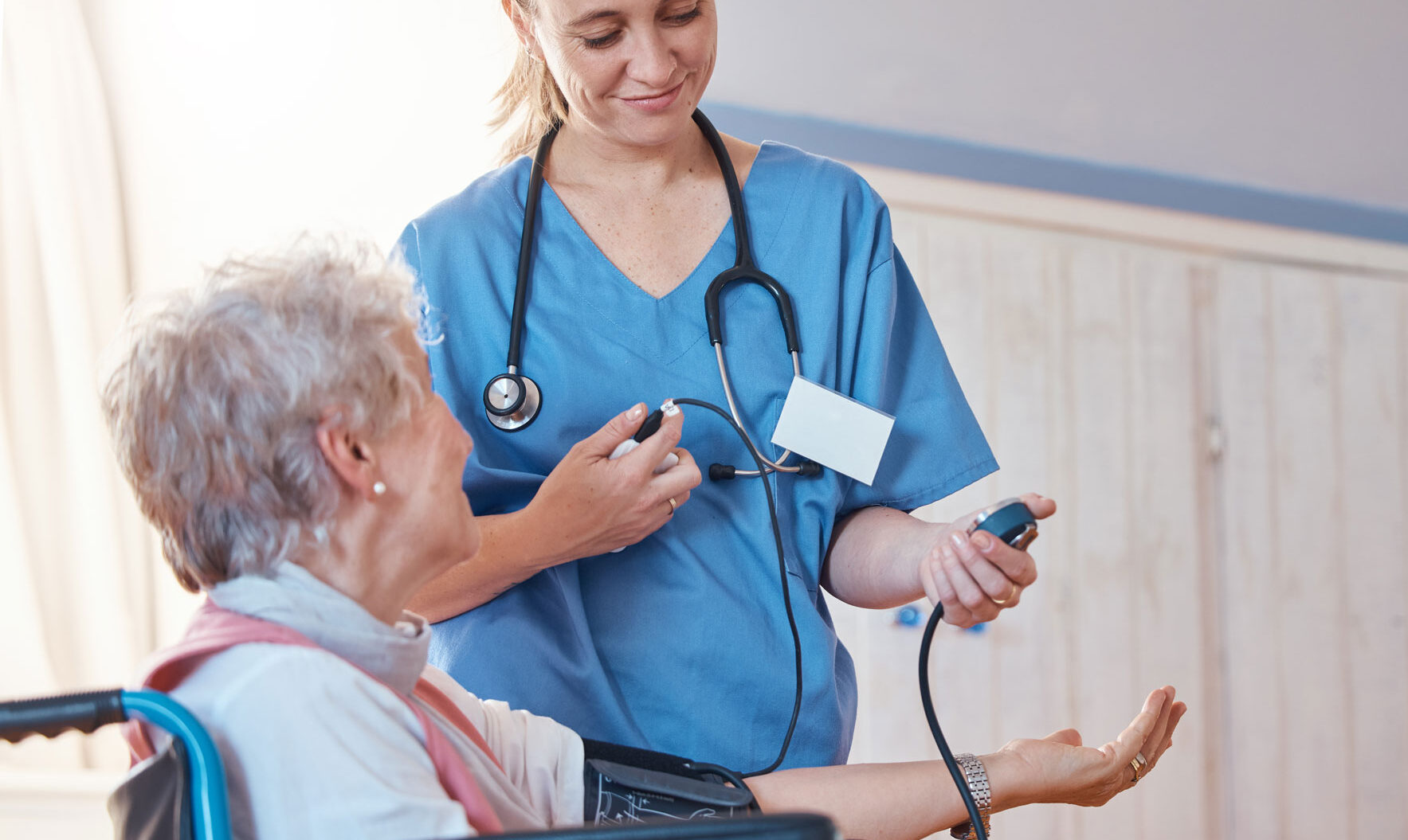 Enfermera de cuidados paliativos con bata azul tomando la presión arterial de una anciana en silla de ruedas