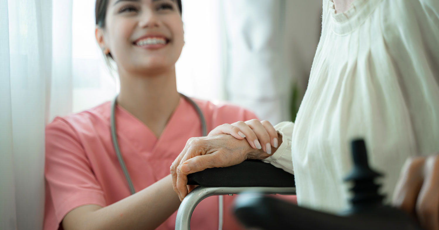 Image of a hospice care nurse in pink scrubs holding the hand and smiling up at an elderly in a wheelchair