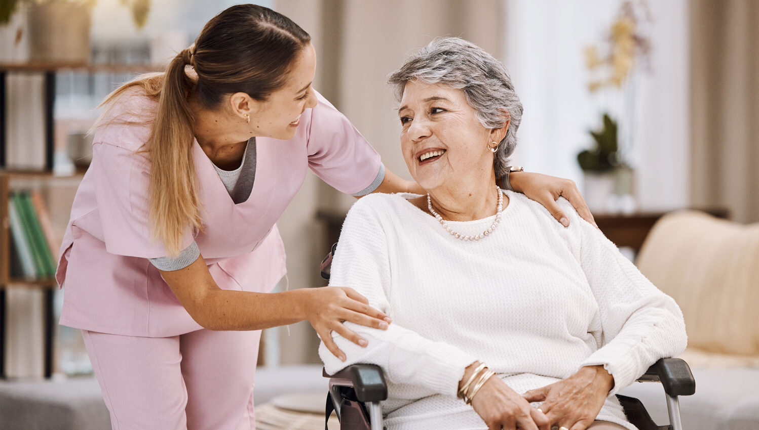 Enfermera de atención domiciliaria vestida de rosa ayudando a un paciente anciano en silla de ruedas