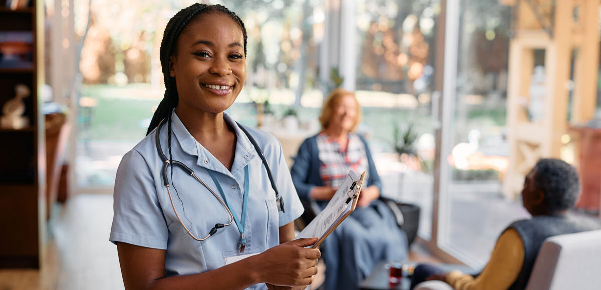 Image of a hospice care nurse smiling and two hospice care elderly in wheelchairs interacting in the background