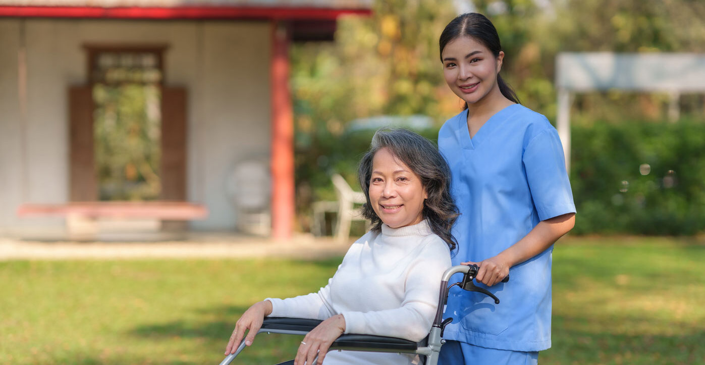 Image of a hospice care nurse in blue scrubs smiling and pushing a hospice care patient in a wheelchair outdoors.