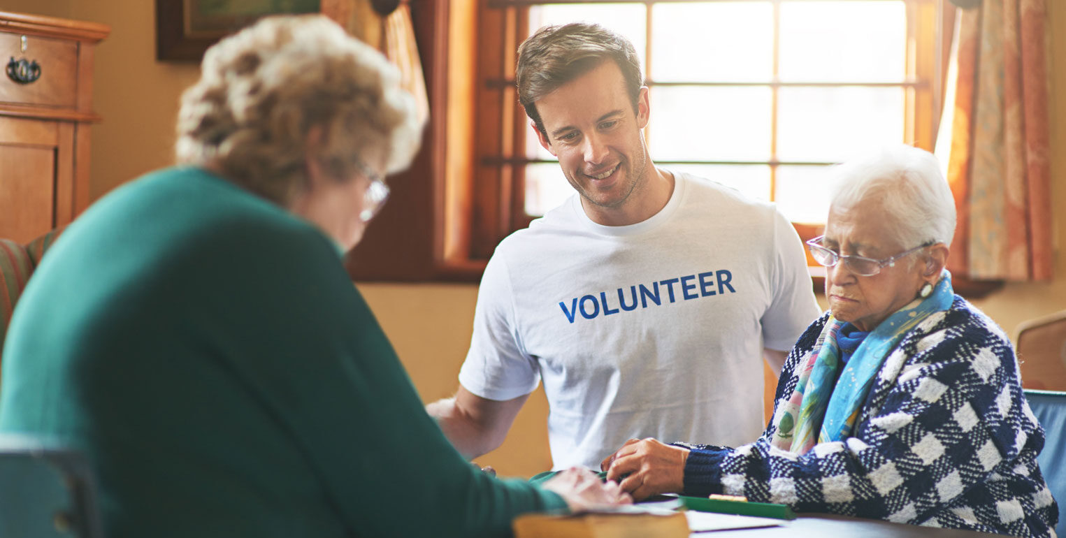 Imagen de un hombre joven con una camisa blanca de voluntario interactuando con dos pacientes ancianos de cuidados paliativos