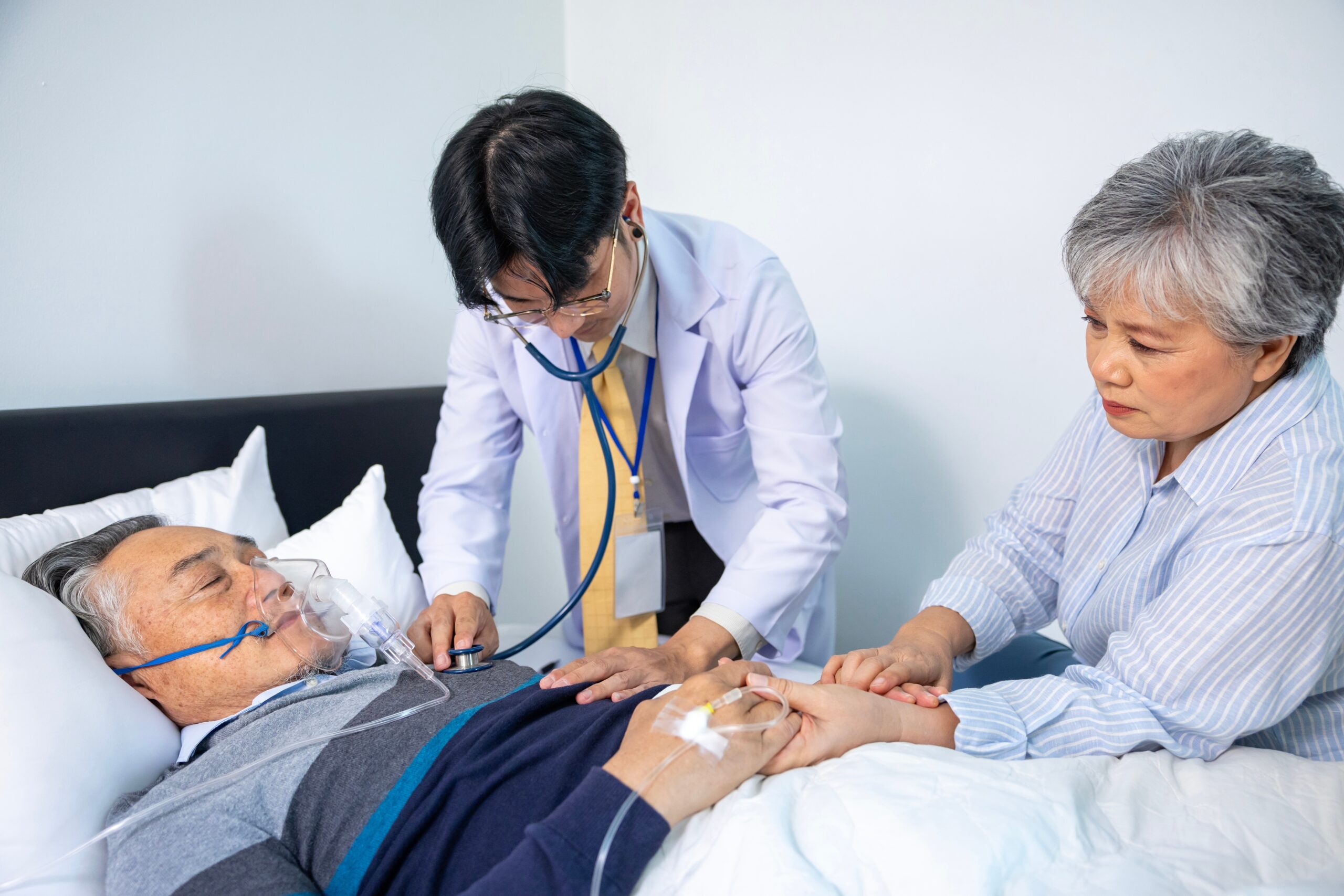 Elderly male patient in bed being comforted by his wife as he is examined by his young male doctor 