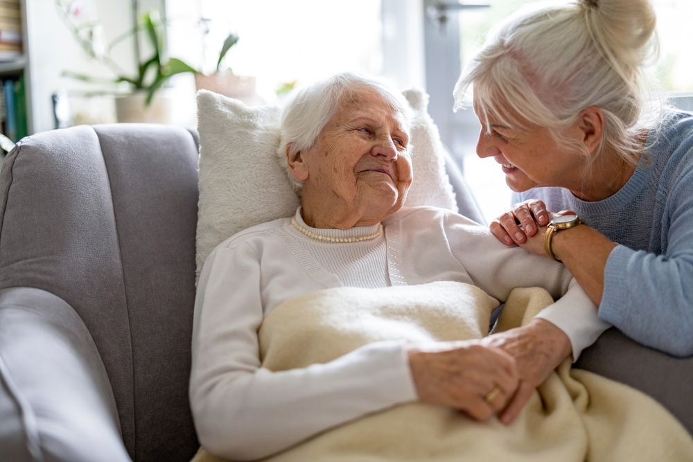 Middle-aged daughter smiling at elderly mother sitting on couch